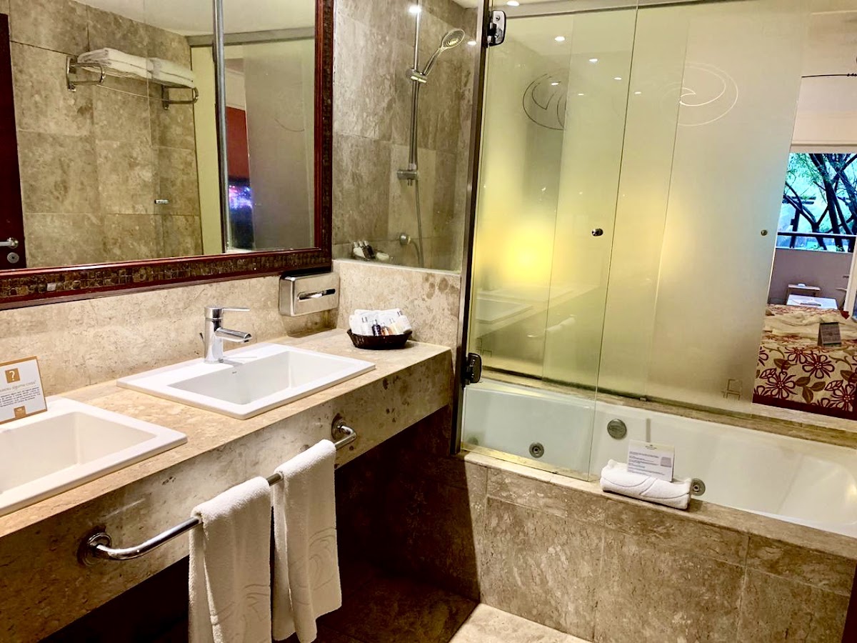 Bathroom with double sink vanity, glass-enclosed shower, and soaking tub with travertine tile walls and dark wood accents.