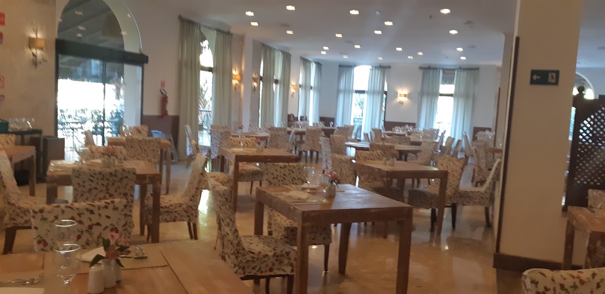 Bright dining room with wooden tables, patterned upholstered chairs, and large windows with curtains overlooking the grounds at Grand Palladium Imbassaí.