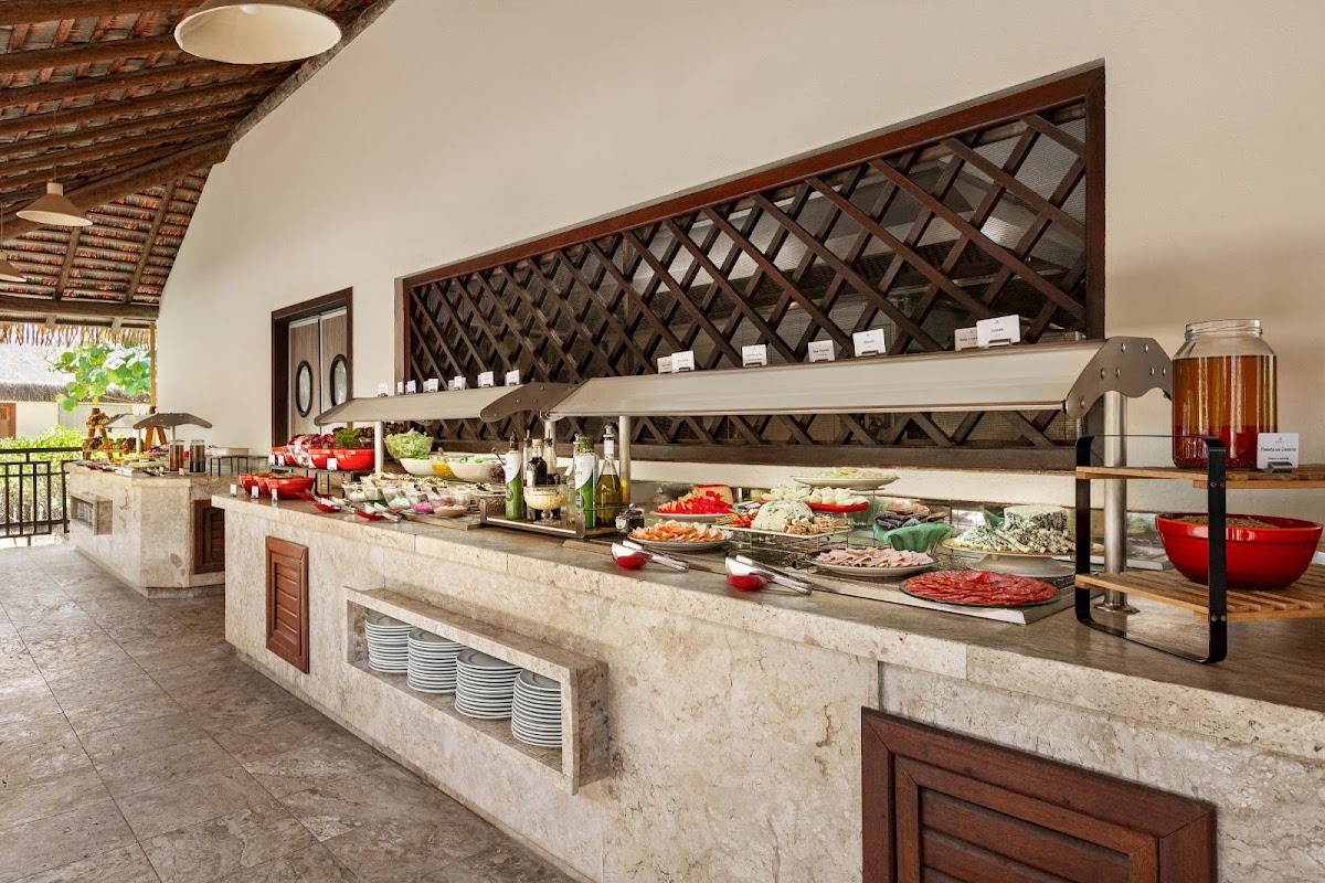 Buffet dining area with stone counters, heat lamps over food stations, and wooden lattice backdrop at Grand Palladium Imbassaí Resort & Spa.