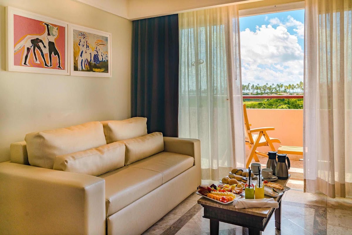 Resort suite with beige sofa, abstract artwork, and glass doors opening to an orange balcony overlooking palm trees and tropical landscape.