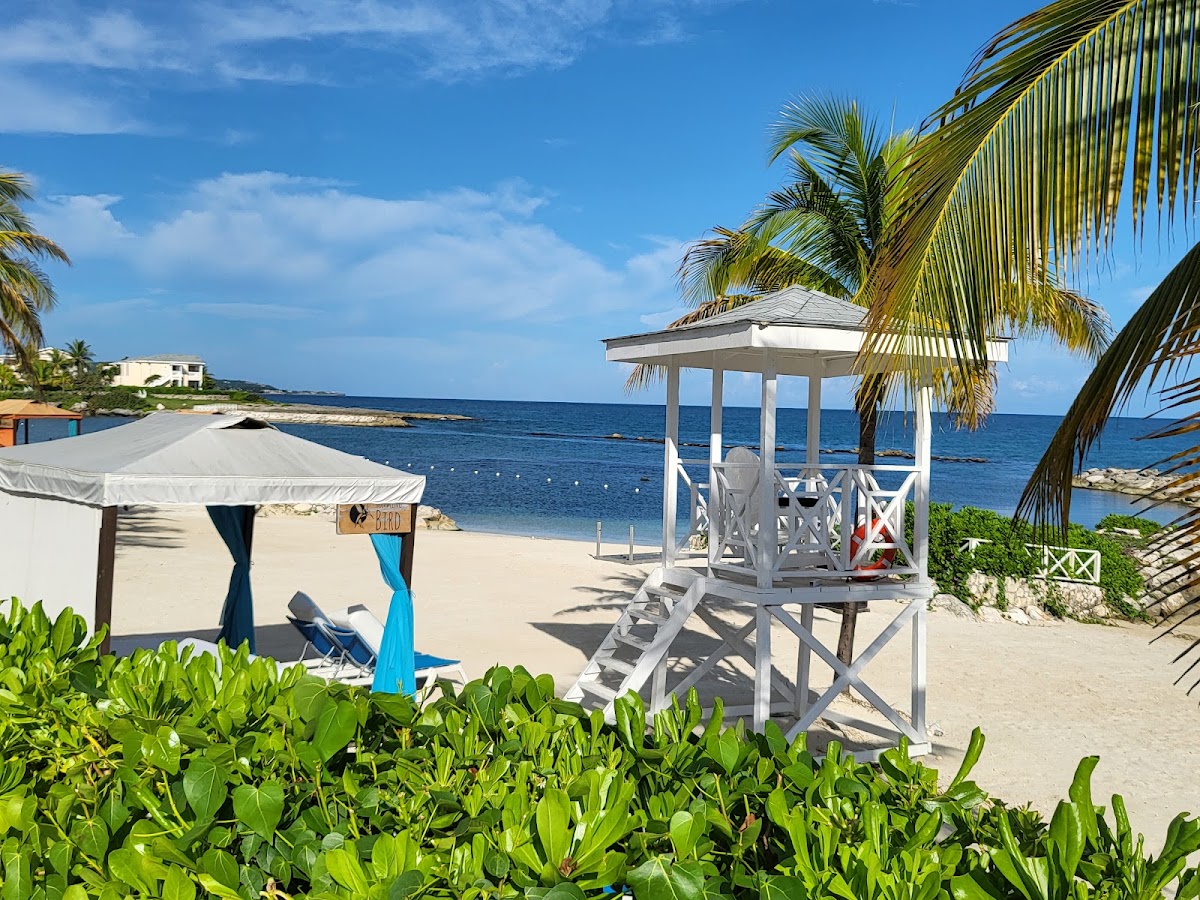 White gazebo and canvas beach cabana overlook the sand and blue ocean at Grand Palladium Jamaica Resort & Spa, framed by palm trees.