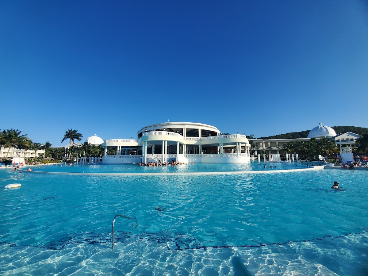 White curved building with blue dome structures sits in bright turquoise pool surrounded by swimmers under clear sky at Grand Palladium Jamaica Resort & Spa.