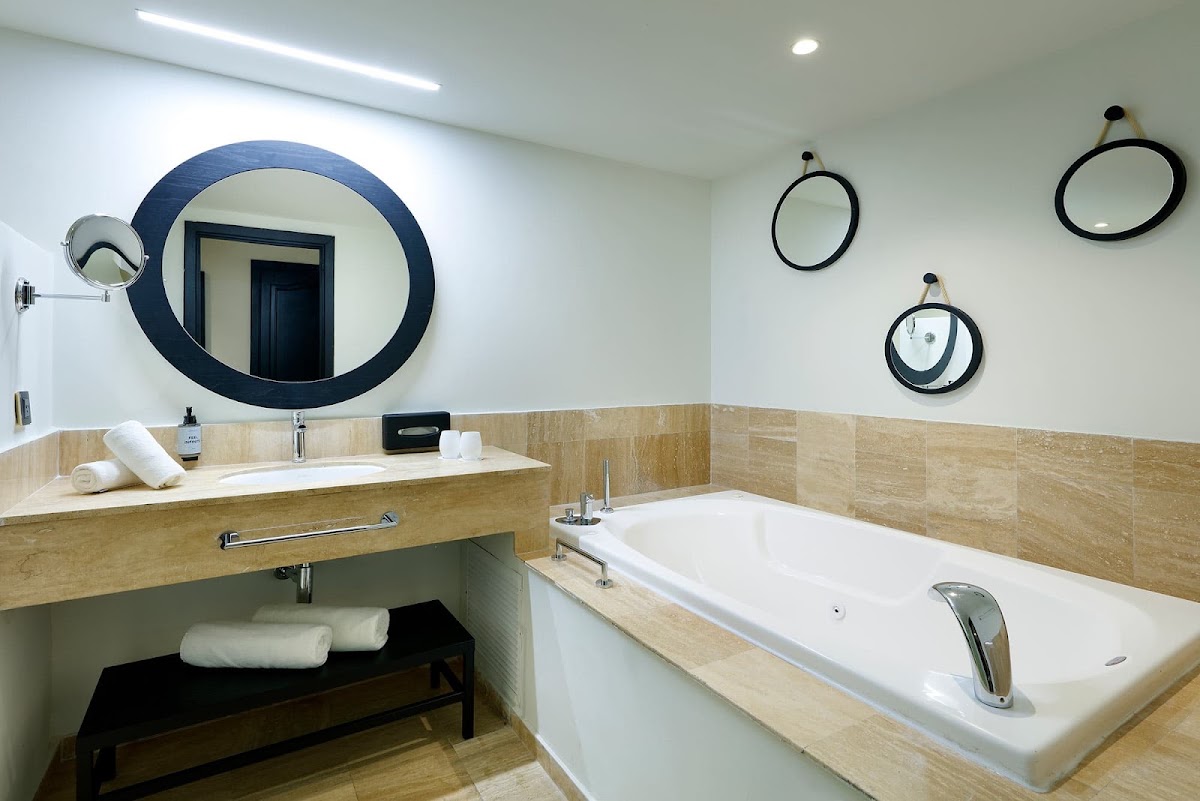 Bathroom with large blue-framed round mirror above sink, white bathtub with tan tile surround, and small circular mirrors on wall.