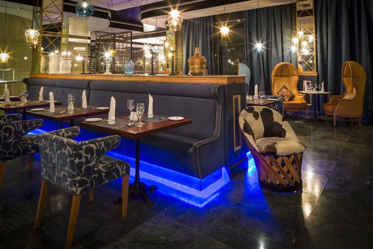 Upscale dining room with blue-lit banquette seating, patterned chairs, wooden tables set with glassware, and hanging pendant lights above a bar.