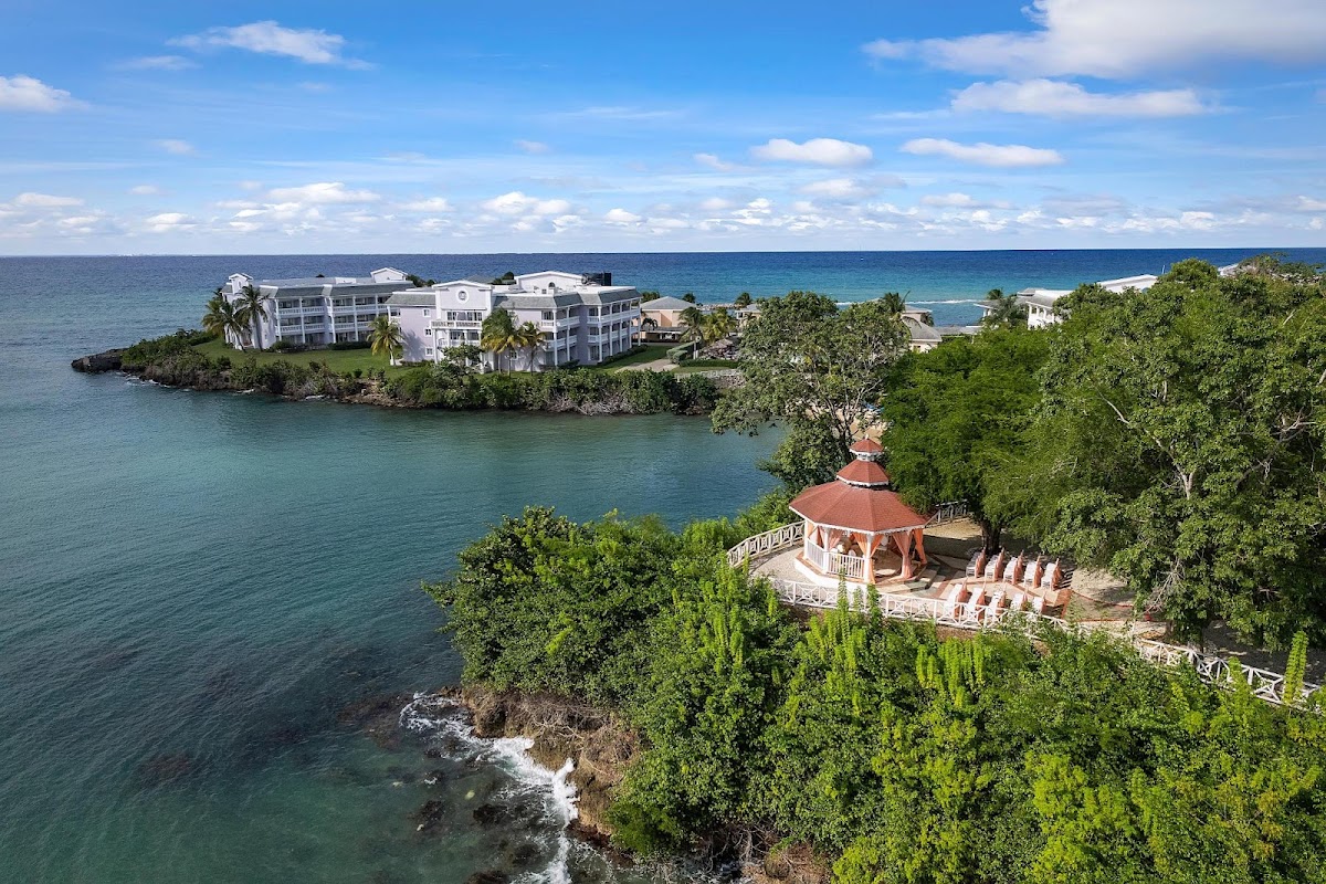 White resort buildings sit on a rocky coastline surrounded by turquoise water and lush vegetation, with a gazebo and lounge chairs on a cliff terrace below.