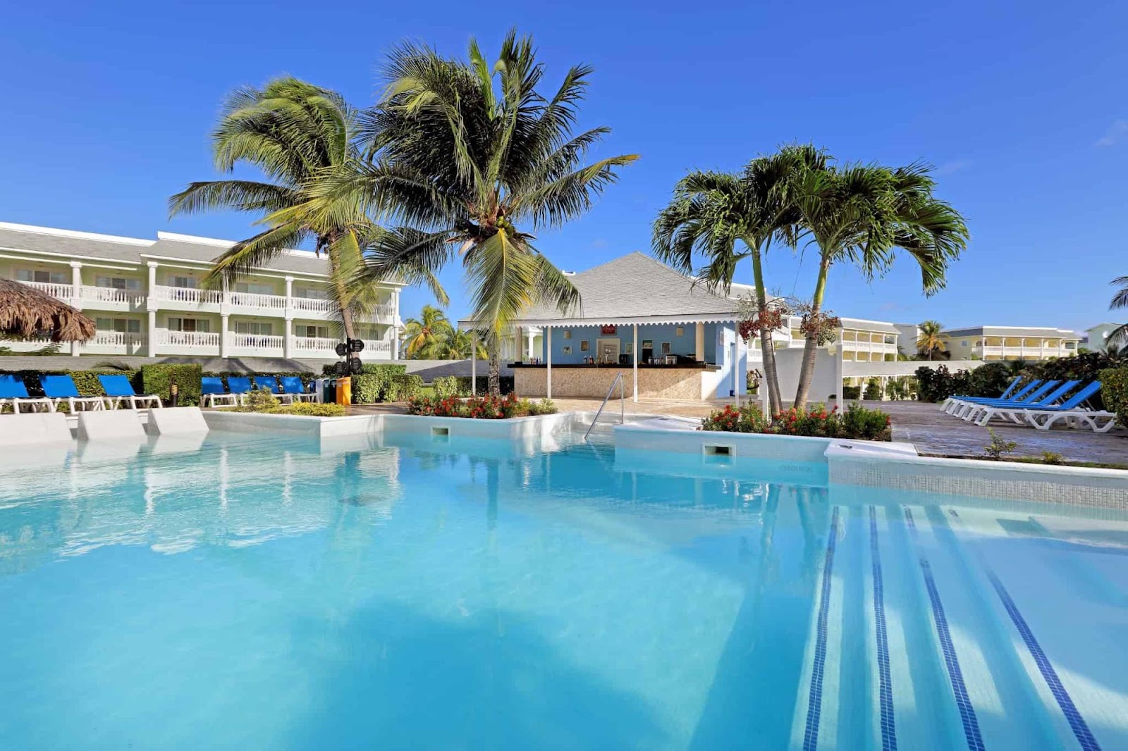 Large turquoise pool with lane markings at Grand Palladium Lady Hamilton Resort & Spa, flanked by white buildings, palm trees, and blue lounge chairs under clear sky.