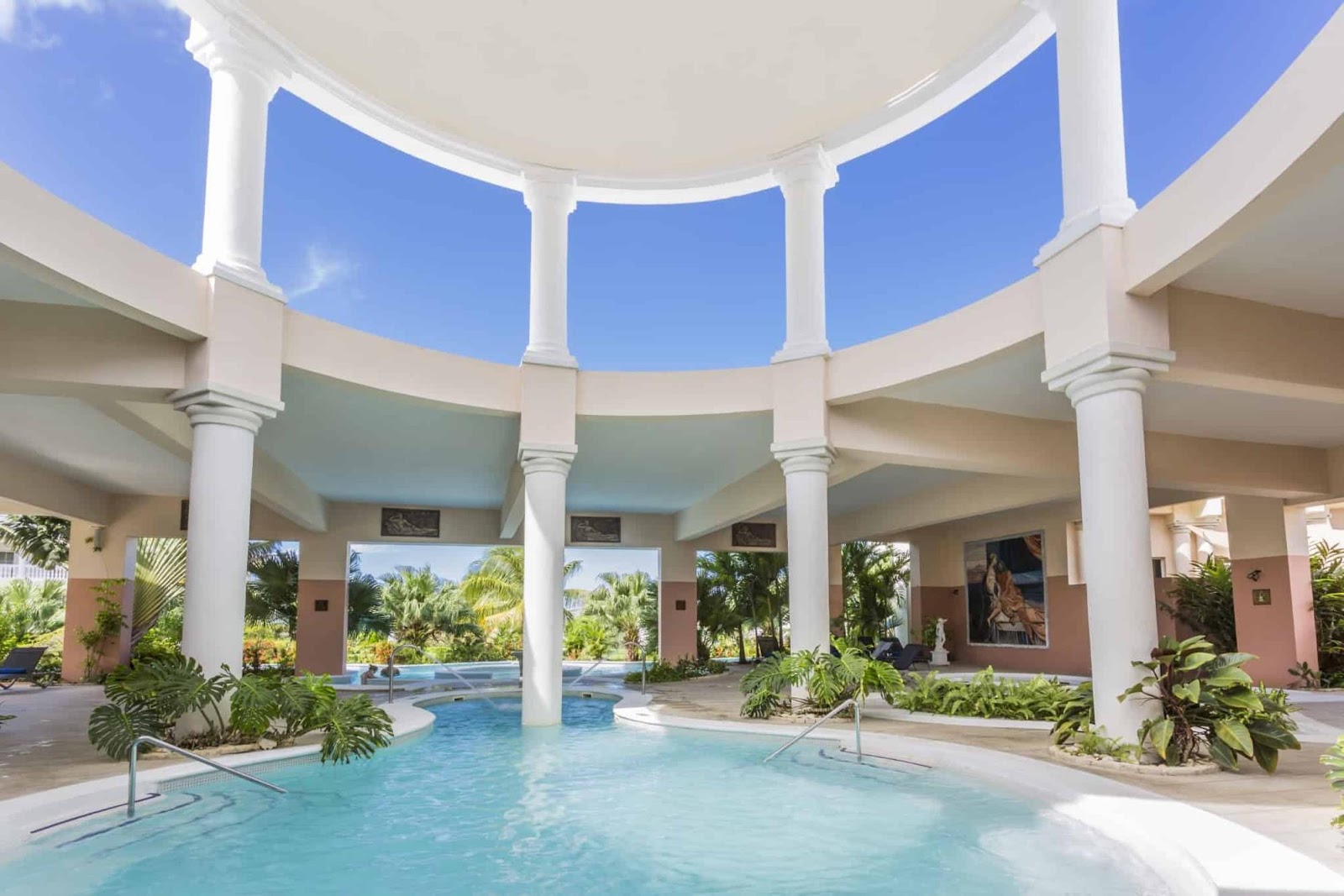 Grand Palladium Lady Hamilton Resort & Spa indoor pool under a curved white colonnade with tropical plants and clear blue sky above.