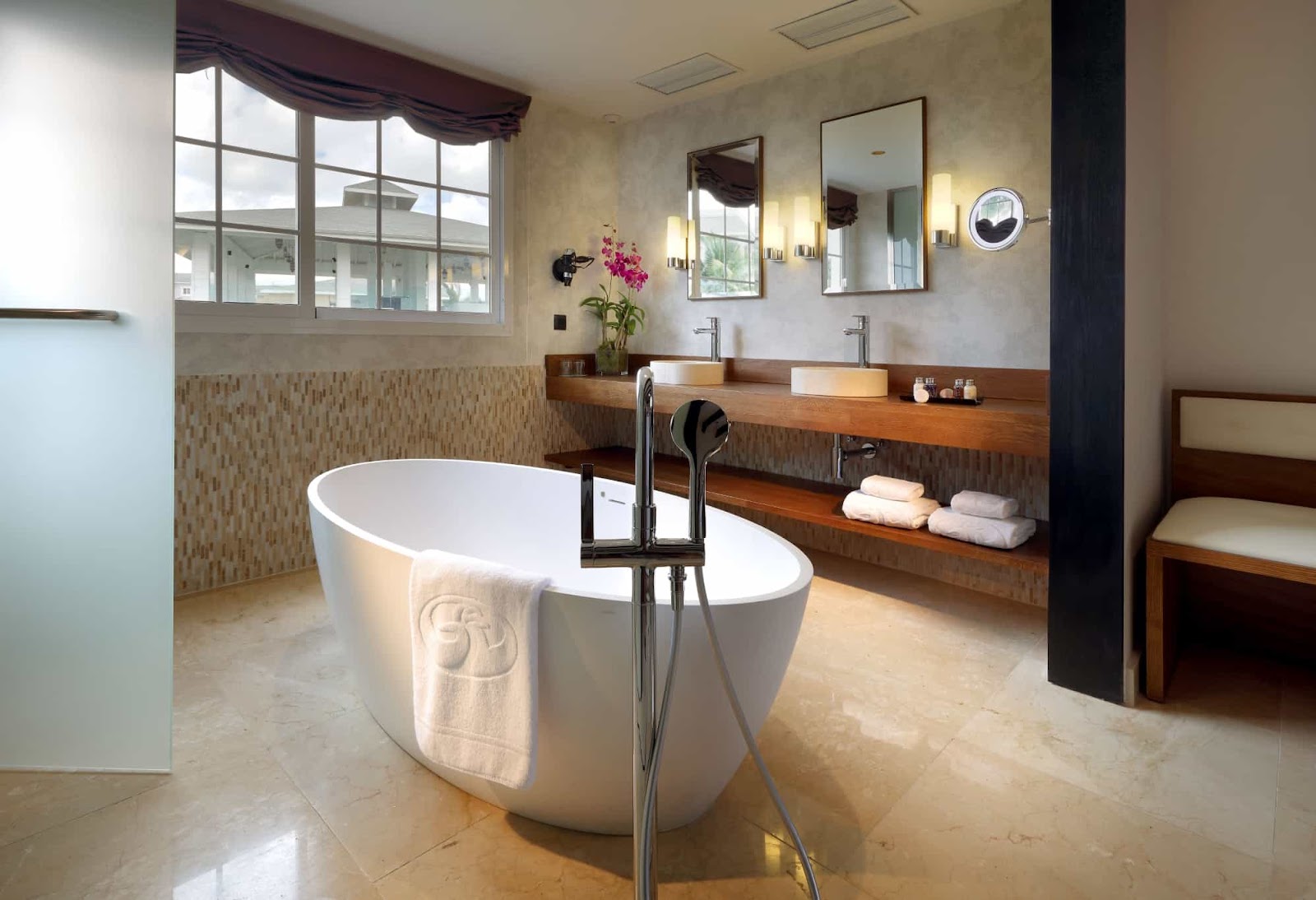 Spa bathroom with freestanding white soaking tub, wooden vanity with dual sinks, pink flowers, and large windows overlooking the resort grounds.