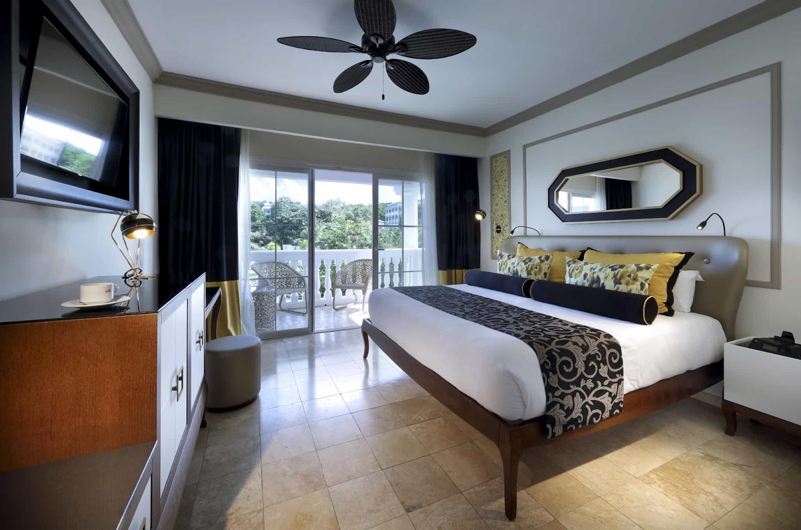 Resort bedroom with king bed, yellow and black accents, ceiling fan, TV, and balcony overlooking tropical grounds.