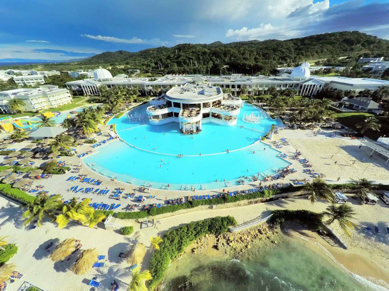 Grand Palladium Lady Hamilton Resort & Spa's circular lagoon pool surrounded by loungers, beach, and white buildings against forested hills.