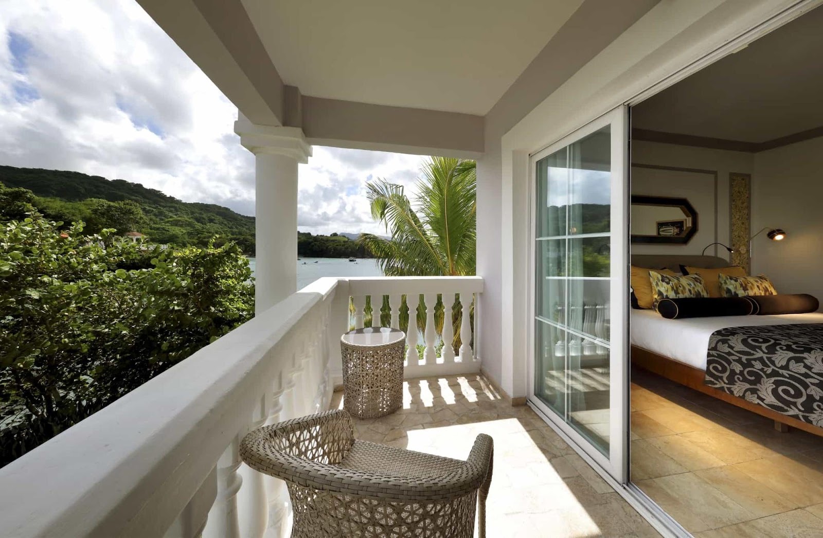 Resort room balcony with woven chair overlooking tropical bay, palm fronds, and green hillside.
