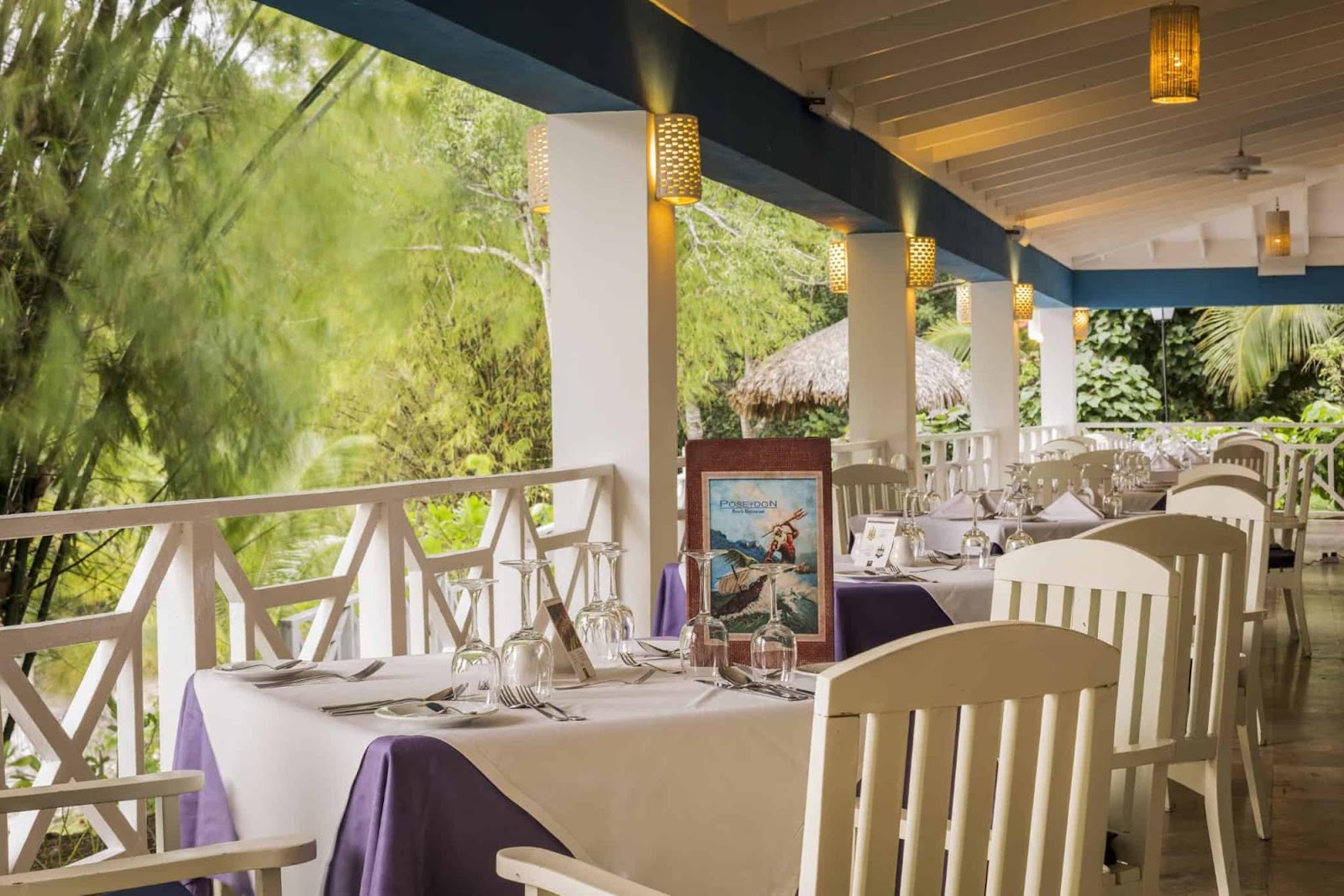 Covered dining area at Grand Palladium Lady Hamilton with set tables, wooden chairs, lush greenery views, and thatched roof structures beyond.