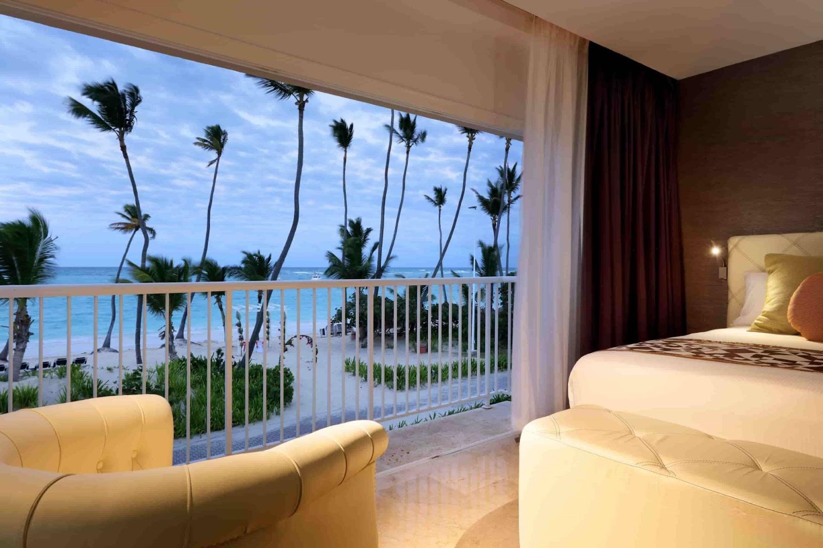 Beachfront resort room with cream leather seating, bed, and a balcony overlooking tall palm trees and turquoise ocean waters.