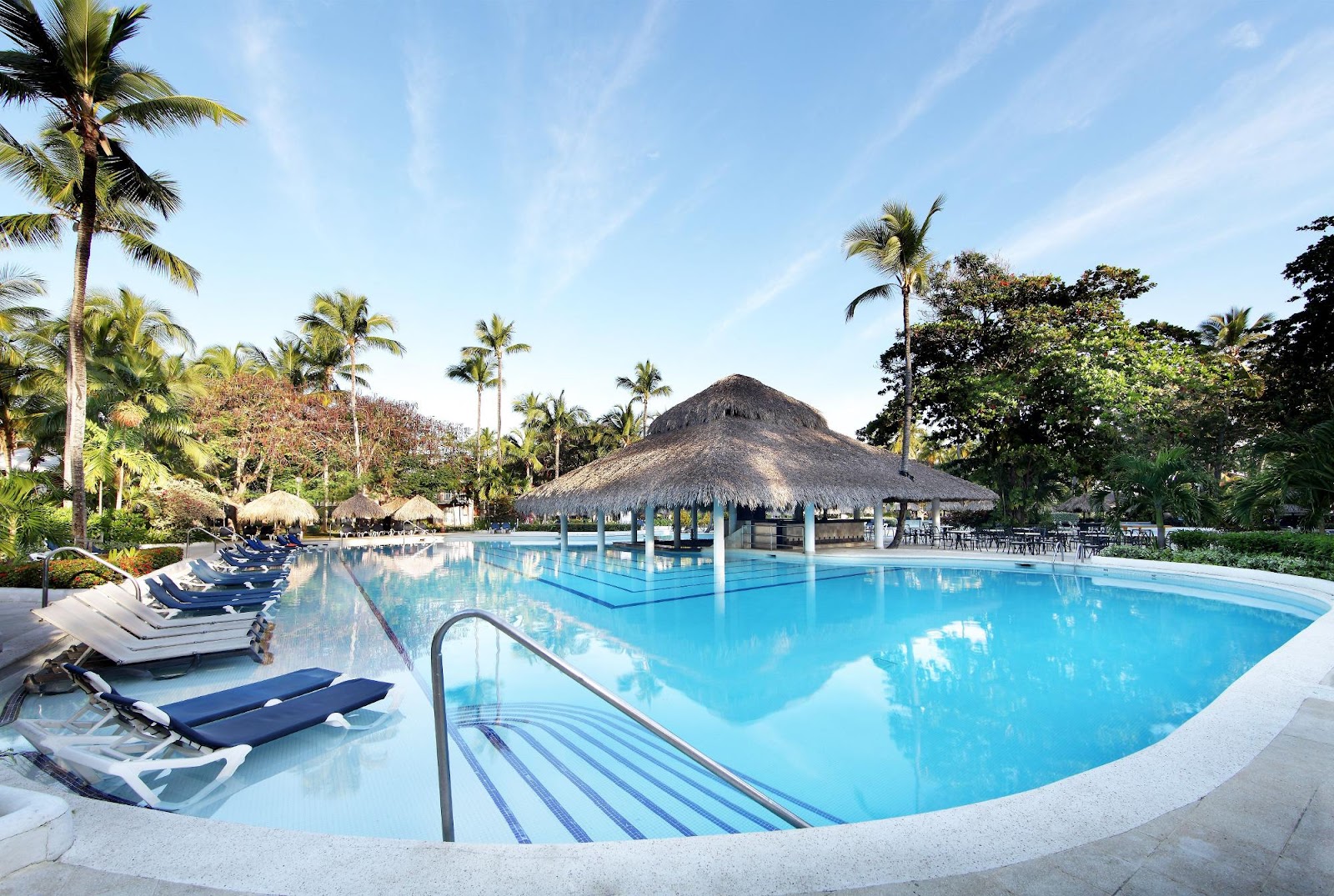 Large resort pool with blue loungers, thatched-roof bar in center, palm trees, and lush tropical grounds under clear sky.