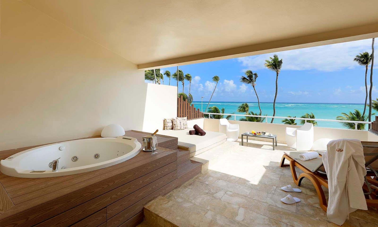 Resort suite with soaking tub, lounge area, and ocean view balcony overlooking turquoise water and palm trees.