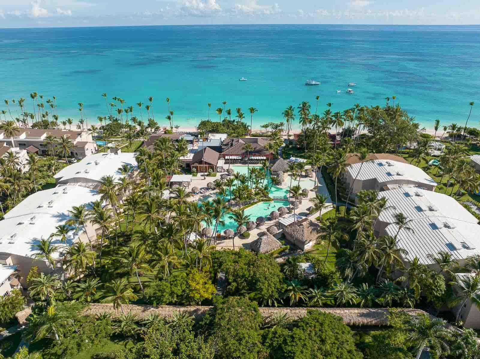 Spacious resort suite with beige sofa, four-poster canopy bed with white curtains, and terrace access overlooking tropical gardens.