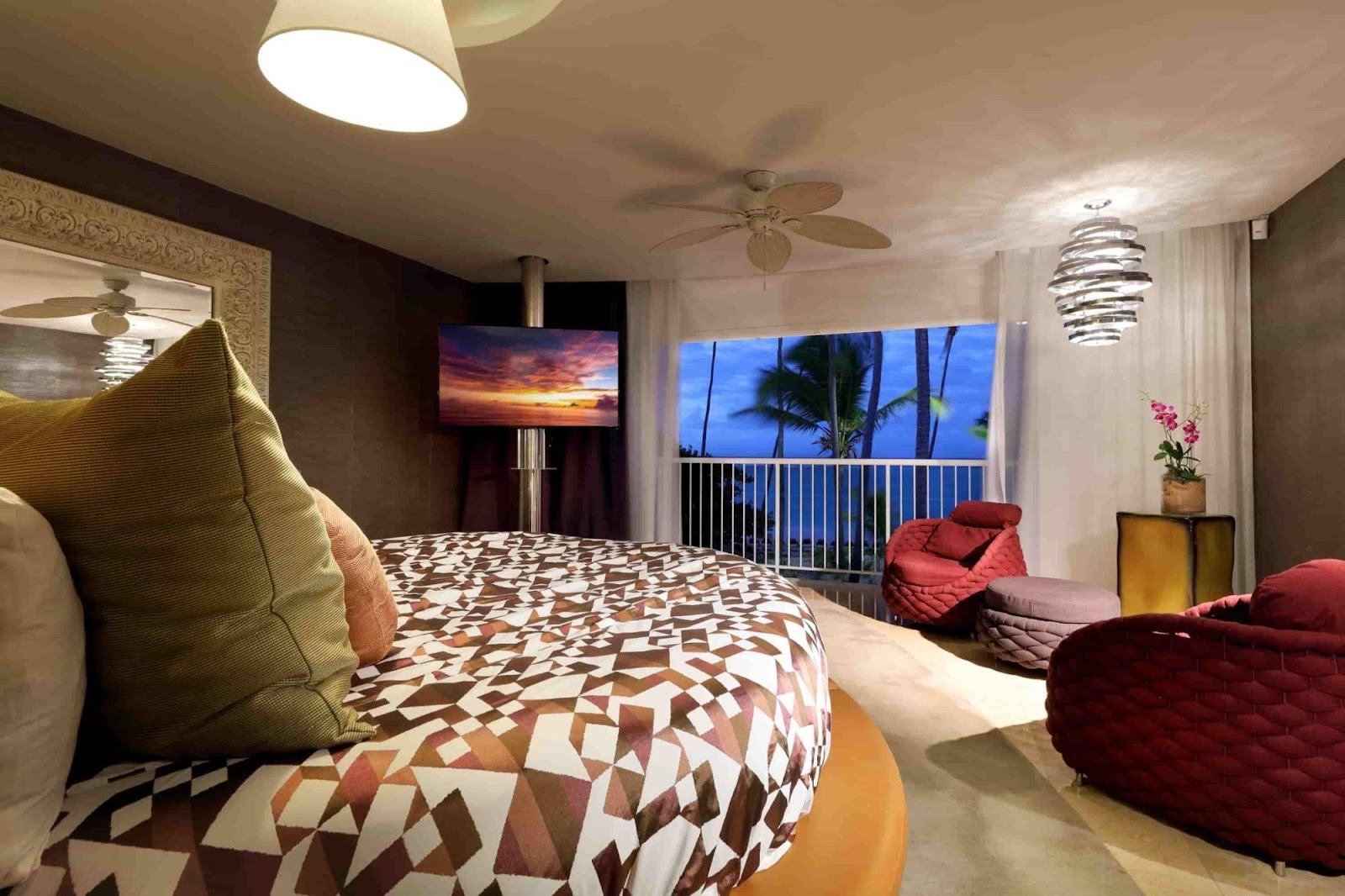 Resort suite with freestanding soaking tub in foreground, dual vanities with vessel sinks, wooden room dividers, bed with ocean-view balcony visible in background.