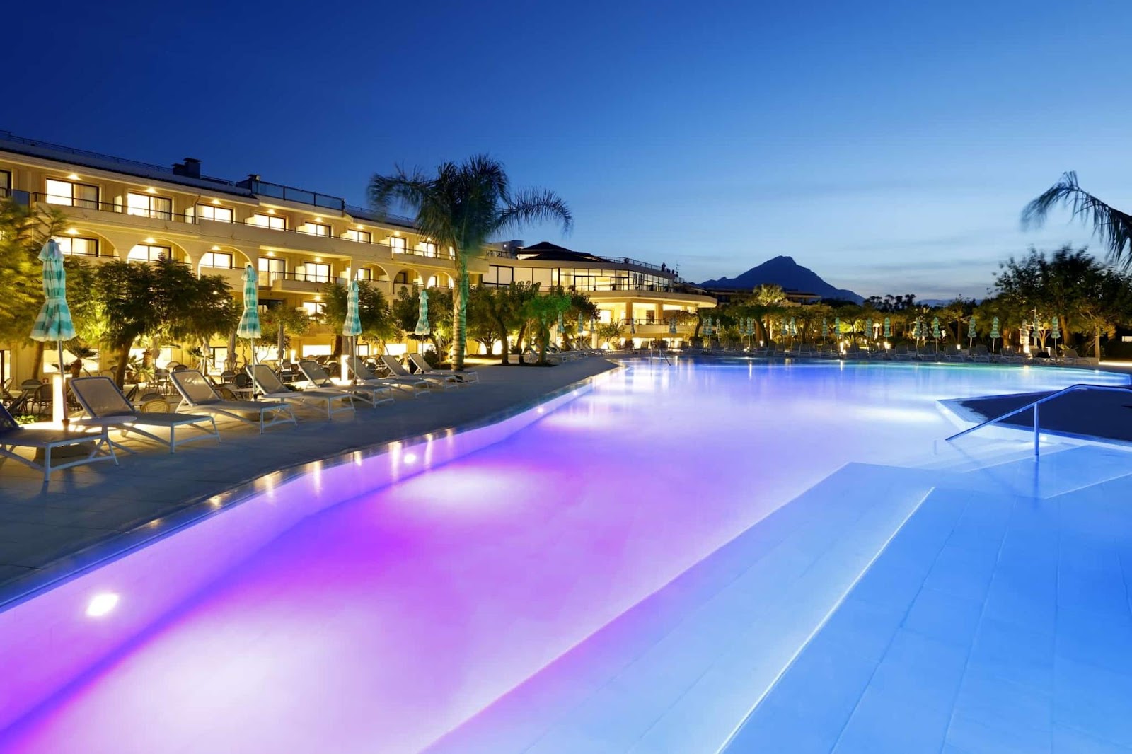 Illuminated pool with purple and blue lighting at dusk, flanked by loungers and palm trees, with a yellow-lit resort building and distant mountain in the background.