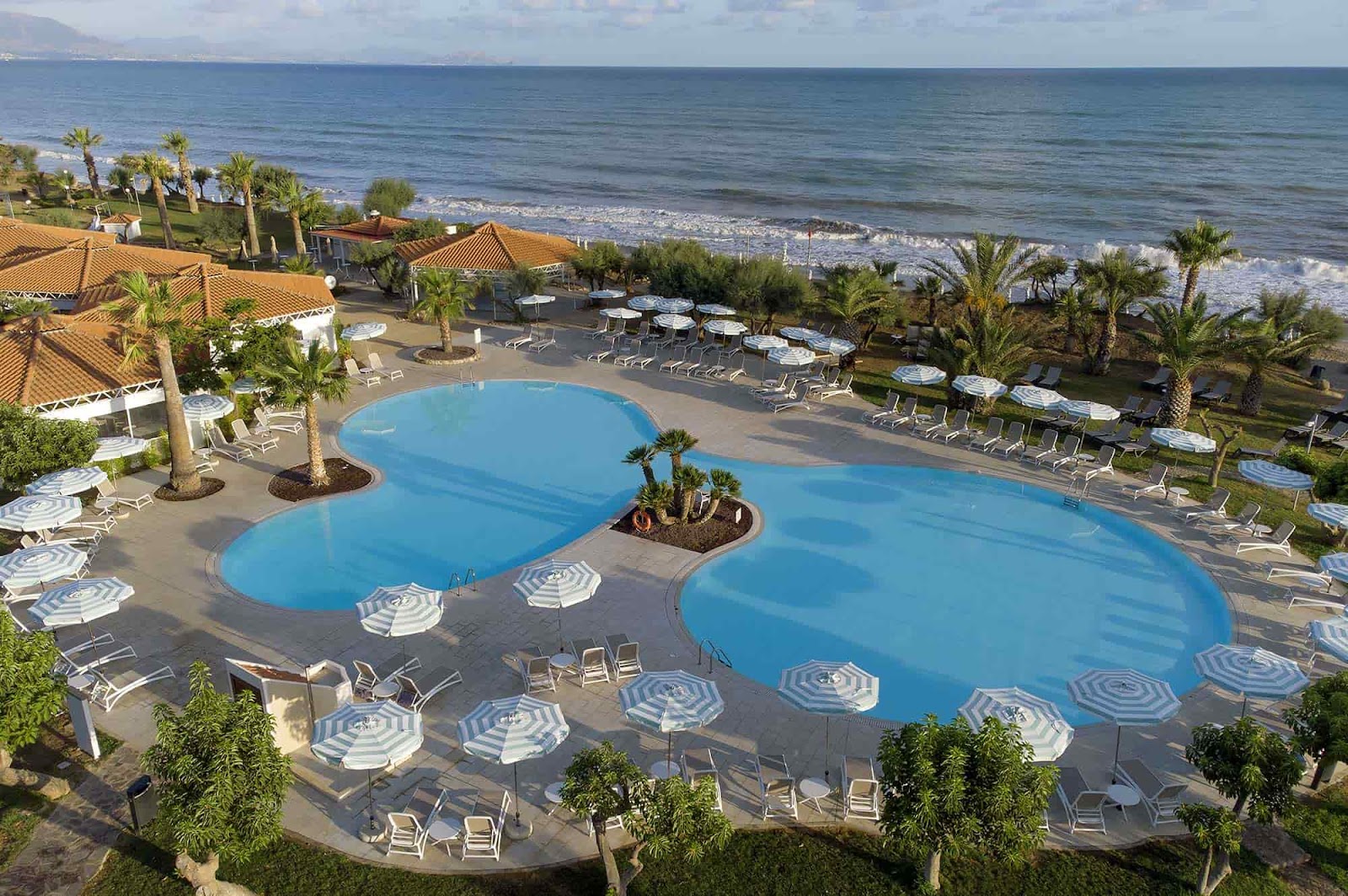 Resort pools with blue umbrellas and loungers overlook a sandy beach and Mediterranean Sea with palm trees and tiled-roof buildings.