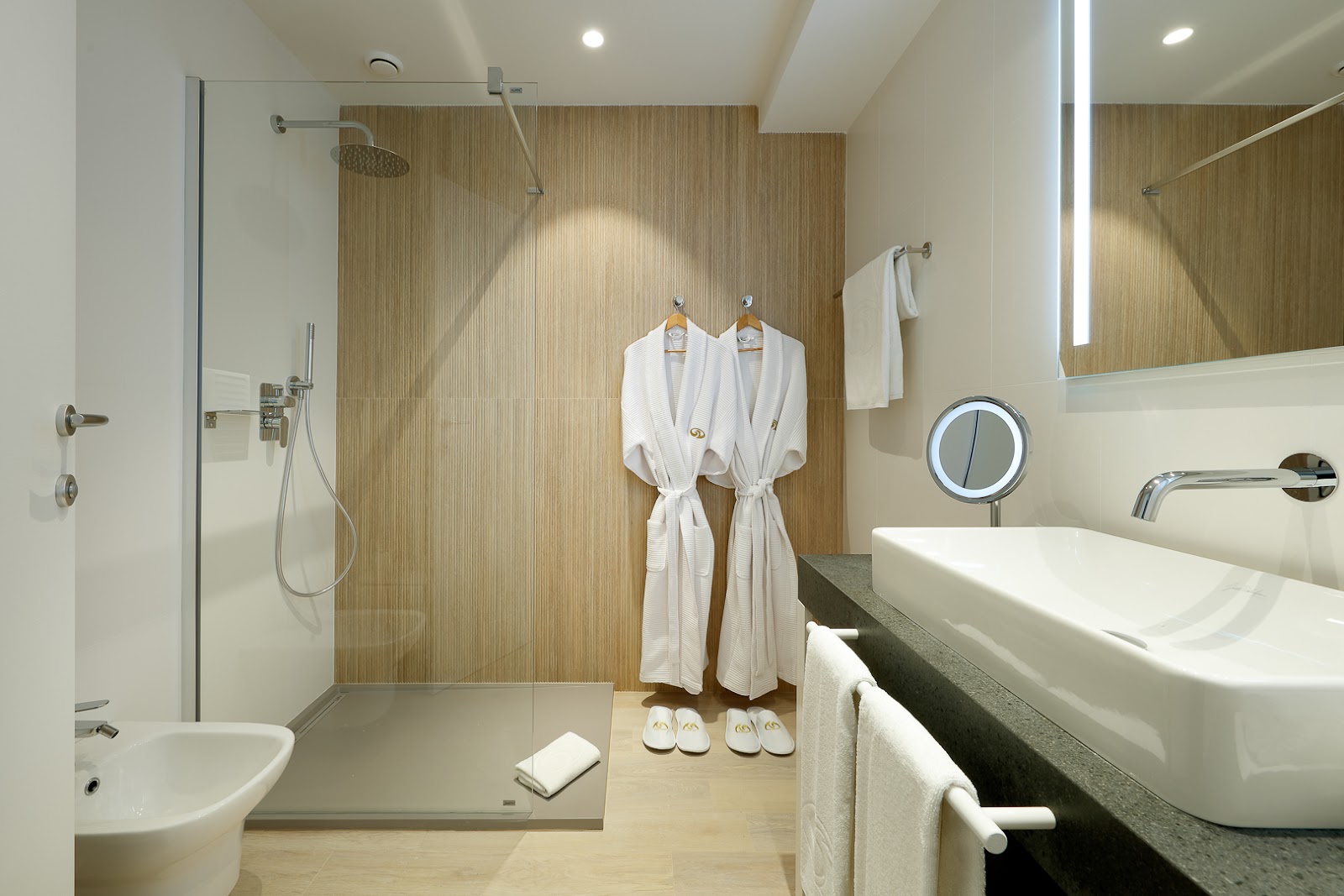 Modern bathroom with walk-in shower, dual white robes on hangers, vessel sinks, and wood-textured accent wall.