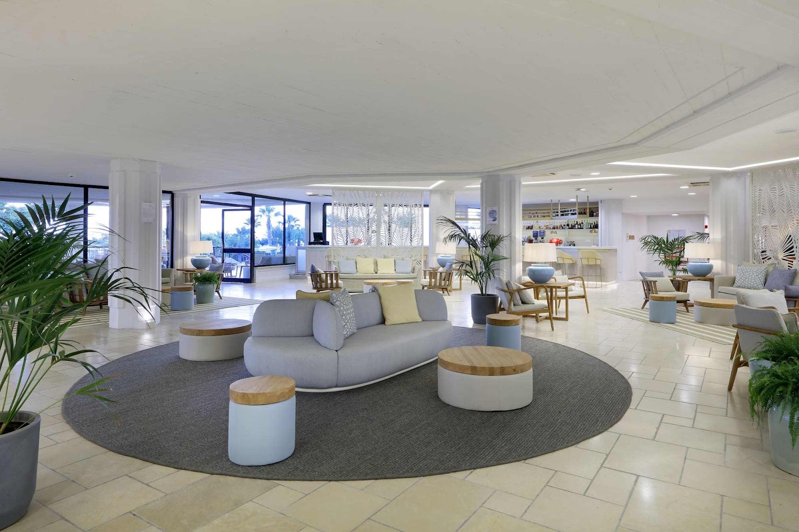 Modern lobby with curved gray sofa, wooden side tables, potted plants, and ocean views through floor-to-ceiling windows at Grand Palladium Sicilia Resort & Spa.