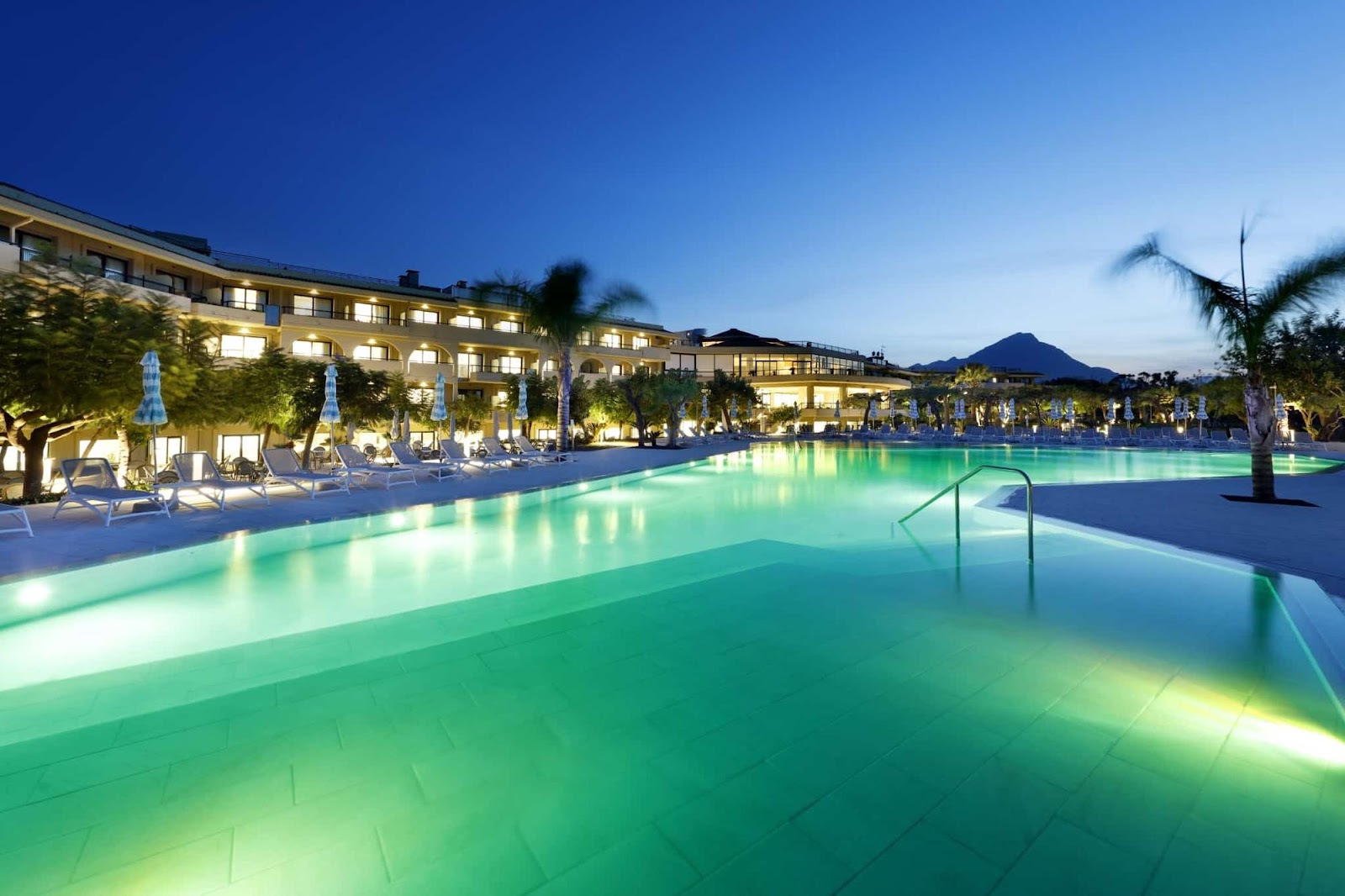Illuminated outdoor pool at dusk with loungers, palm trees, and a multi-story hotel building with lit windows against a mountain backdrop.