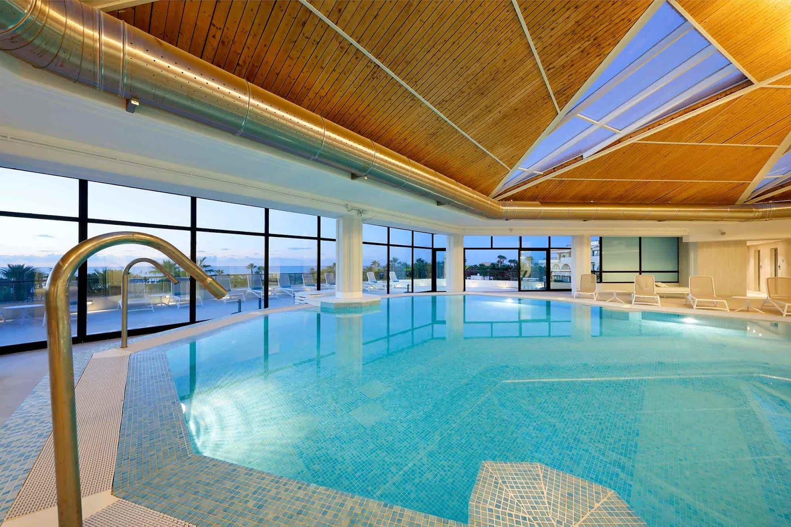 Indoor swimming pool with wooden ceiling and skylights, gold handrail in foreground, lounge chairs along windows overlooking ocean and outdoor pool area.