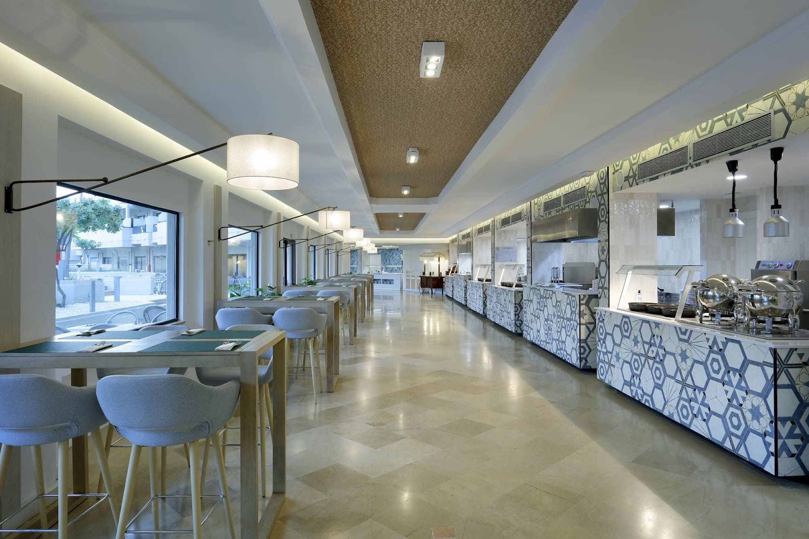 Modern resort buffet restaurant with polished floors, geometric-patterned service counter, pendant lighting, and seating areas overlooking a street.