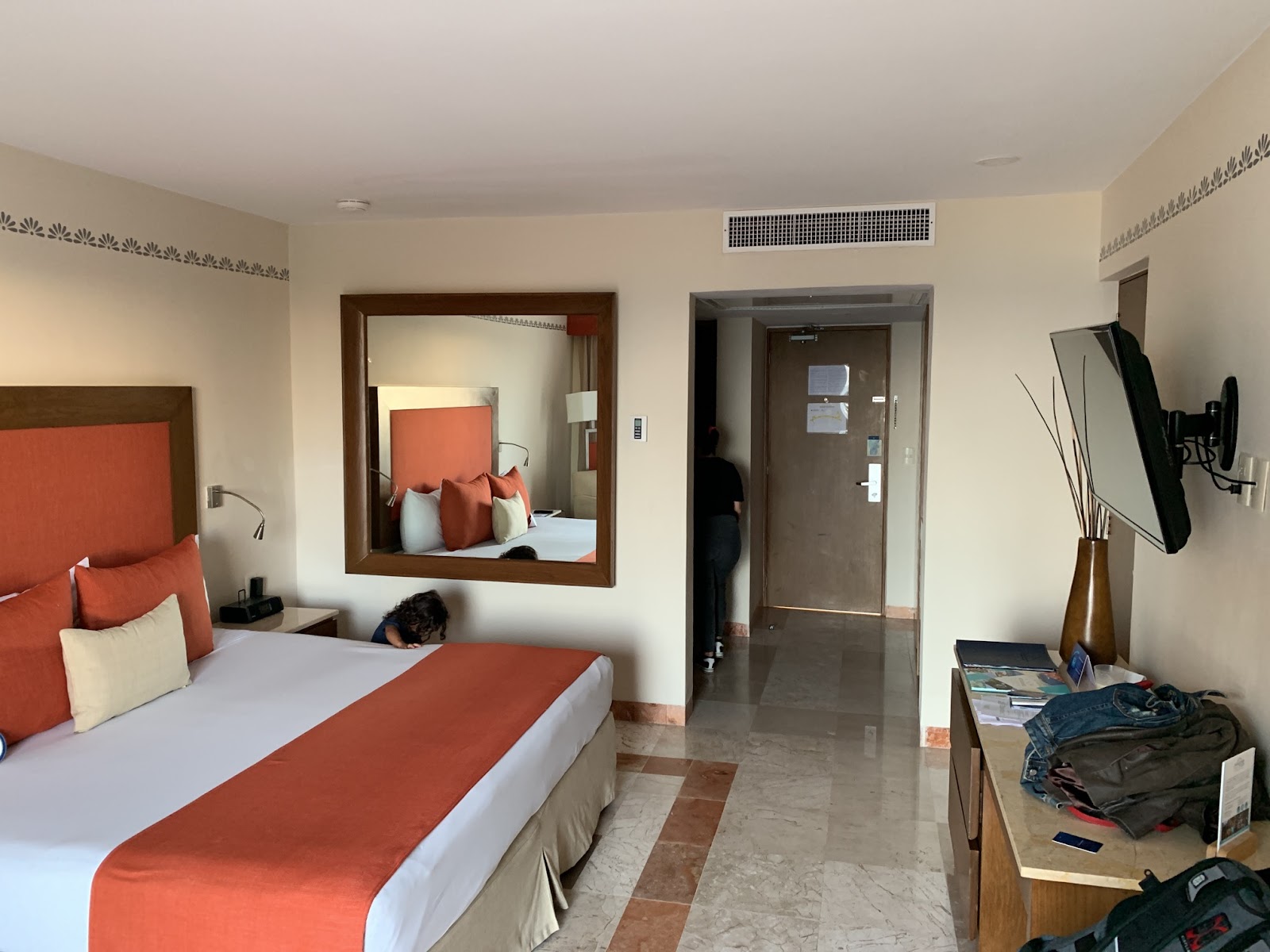 Resort guestroom with queen bed dressed in white linens and orange runner, mirror above bed, and tile flooring with decorative border.