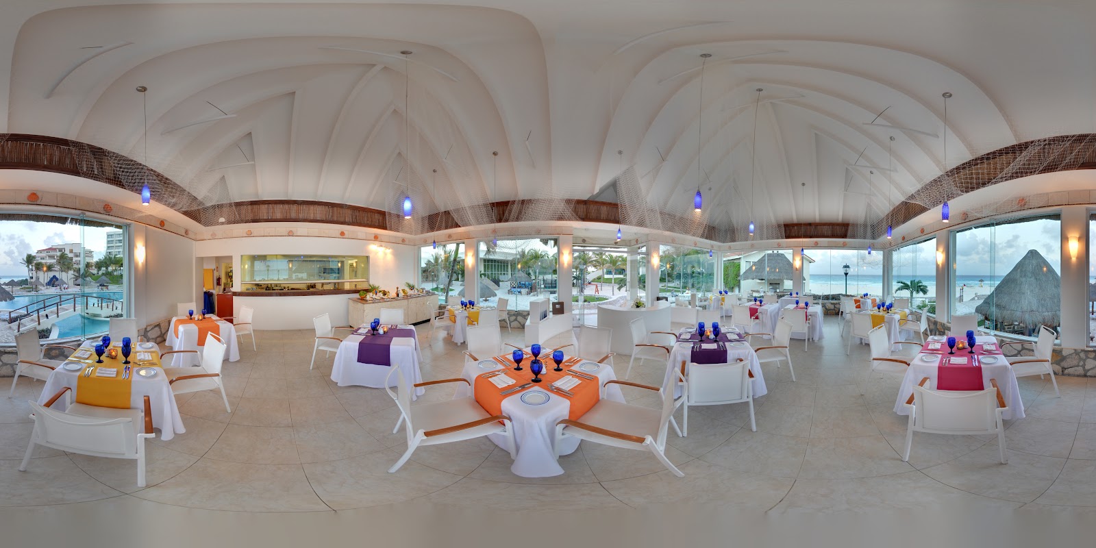 Modern dining hall at Grand Park Royal Cancún with arched white ceiling, round tables dressed in white and colorful linens, ocean and pool views through floor-to-ceiling windows.