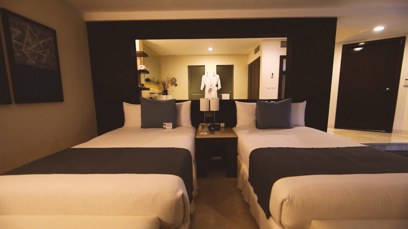 Modern resort hotel room with two queen beds, dark accent wall, vanity with mirror, and bathroom entrance.