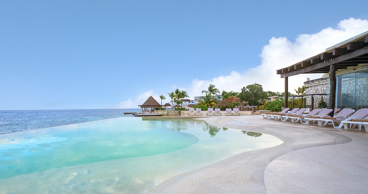 Grand Park Royal Cozumel's pool curves along the sandy beach with lounge chairs, a wooden pergola, and the Caribbean Sea beyond.