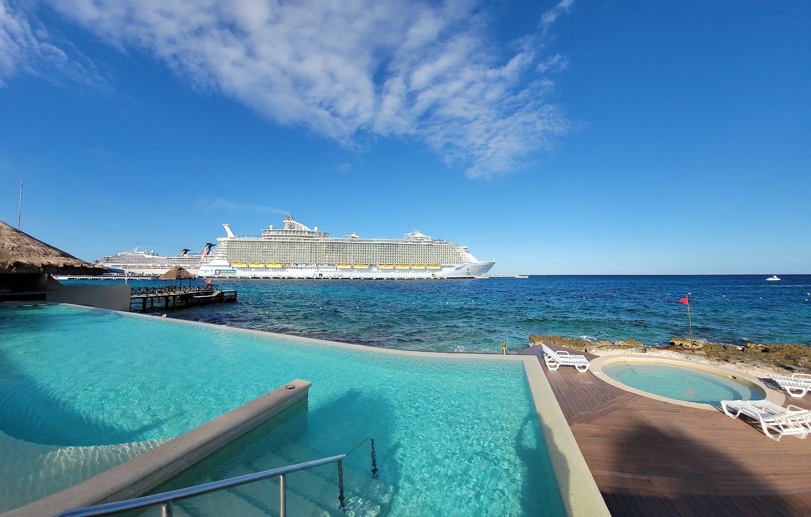 Large cruise ship anchored offshore beyond a resort beach with shallow turquoise water, wooden dock, and lounge chairs.