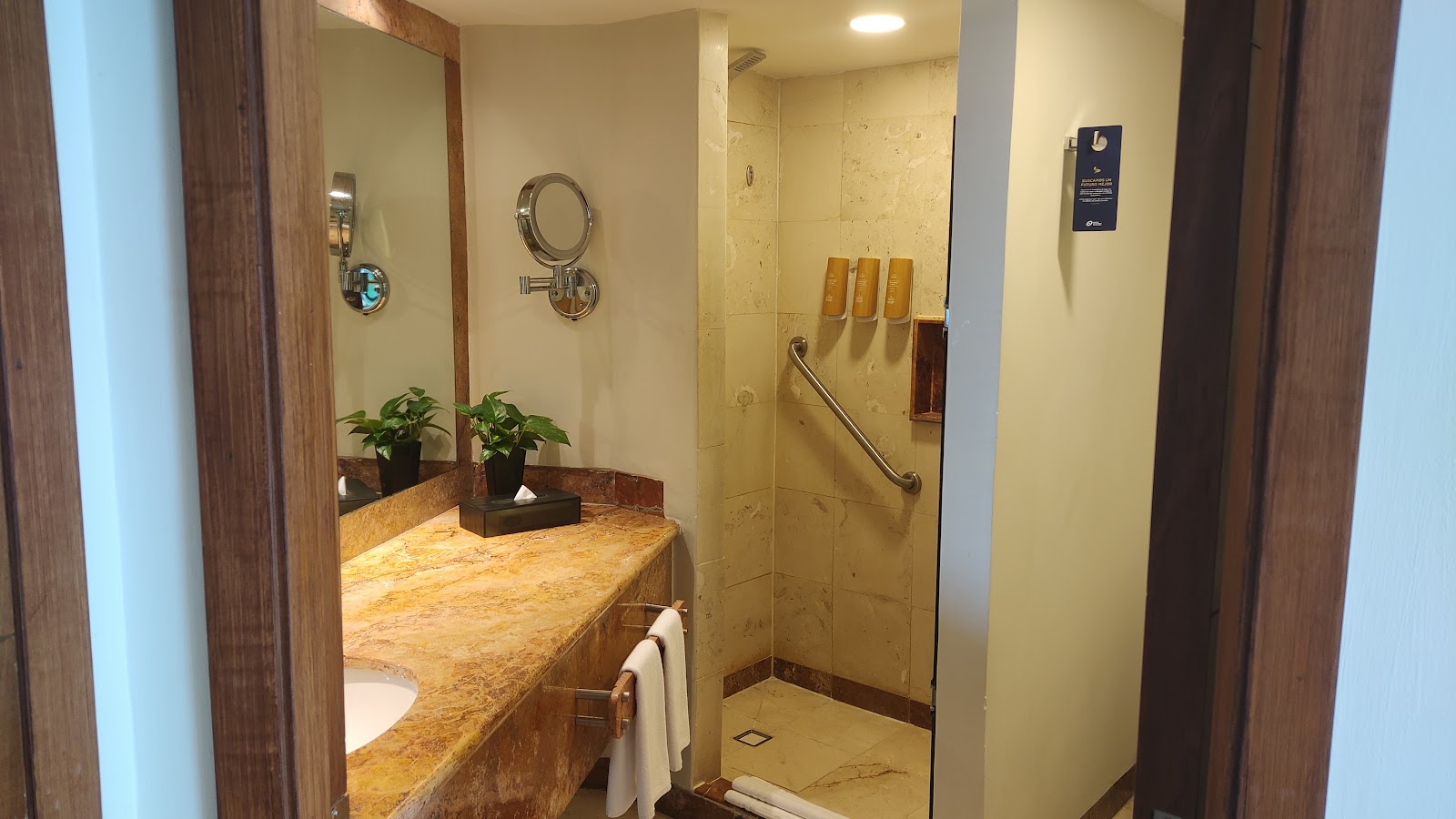 Resort bathroom with marble countertop, dual sink, large mirror, and wooden doors opening to guest room with TV and bed.