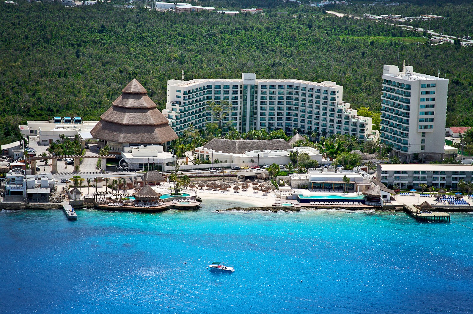Grand Park Royal Cozumel's white multi-story hotel buildings and thatched-roof structures sit on a sandy beach along bright turquoise Caribbean waters, with dense jungle visible behind.