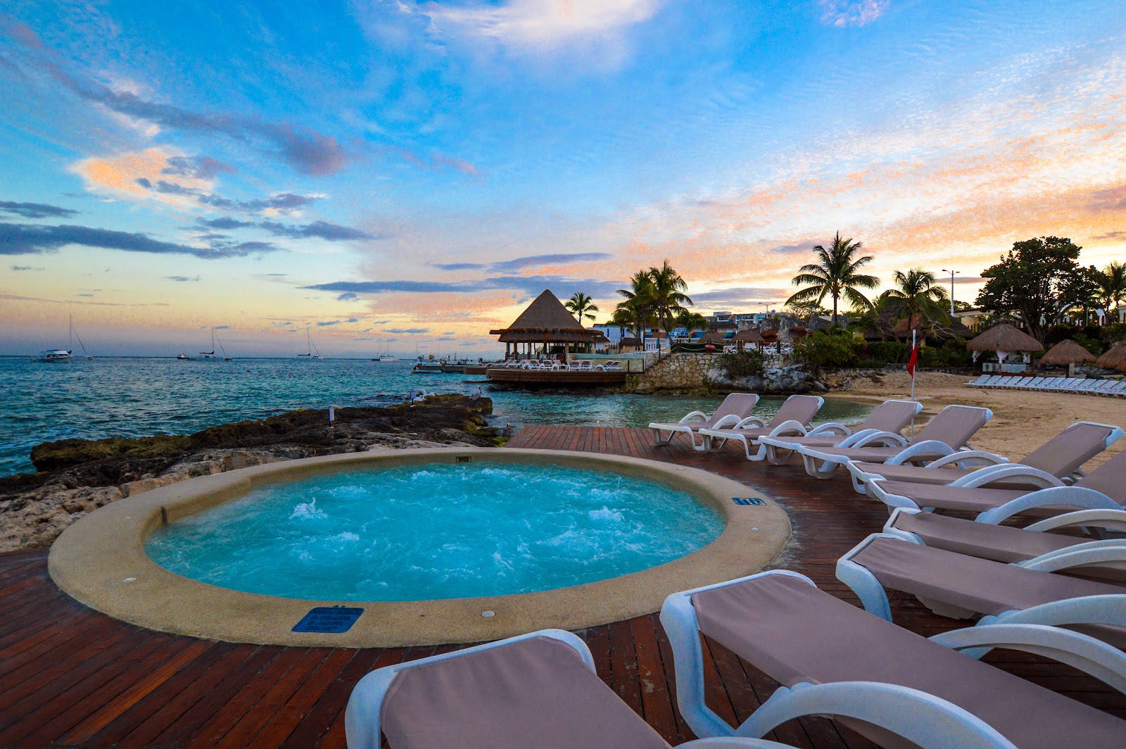 Circular pool on wooden deck with lounge chairs overlooking beach, ocean, and thatched-roof bar at sunset in Cozumel.