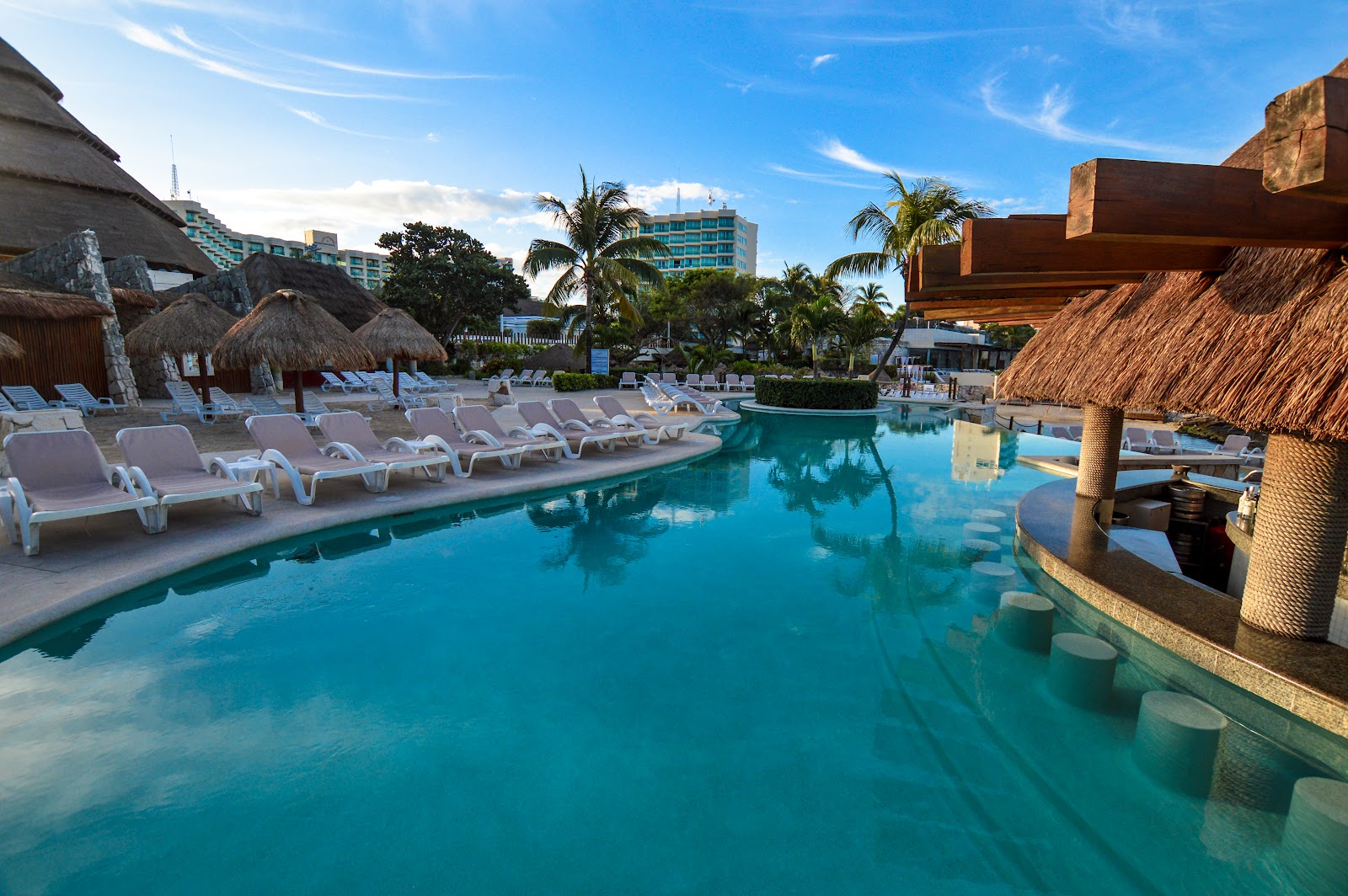 Turquoise pool with white loungers, thatched-roof palapas, and resort buildings under blue sky at Grand Park Royal Cozumel.
