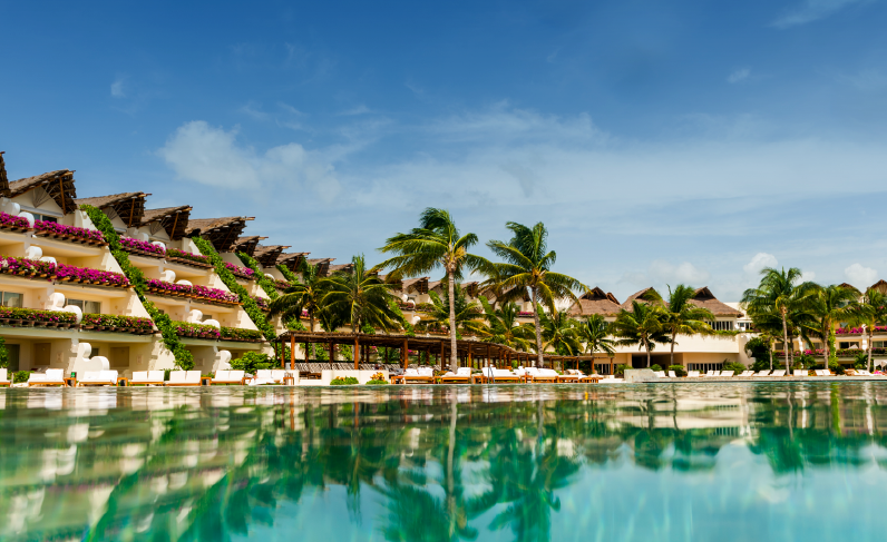 Tiered beachfront buildings with magenta flowers and thatched palapas overlook turquoise water and white sand at Grand Velas Riviera Maya.