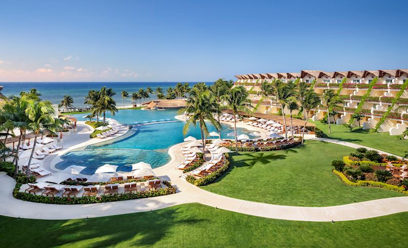 Aerial view of Grand Velas Riviera Maya with curved pool, palm trees, beachfront, and tiered room buildings along the Caribbean shore.