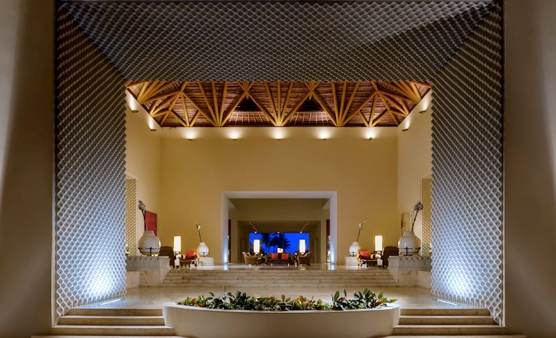 Grand Velas Riviera Maya's lobby features a cream-colored interior with geometric wooden ceiling details, illuminated pendant lights, and a central planter with tropical plants.