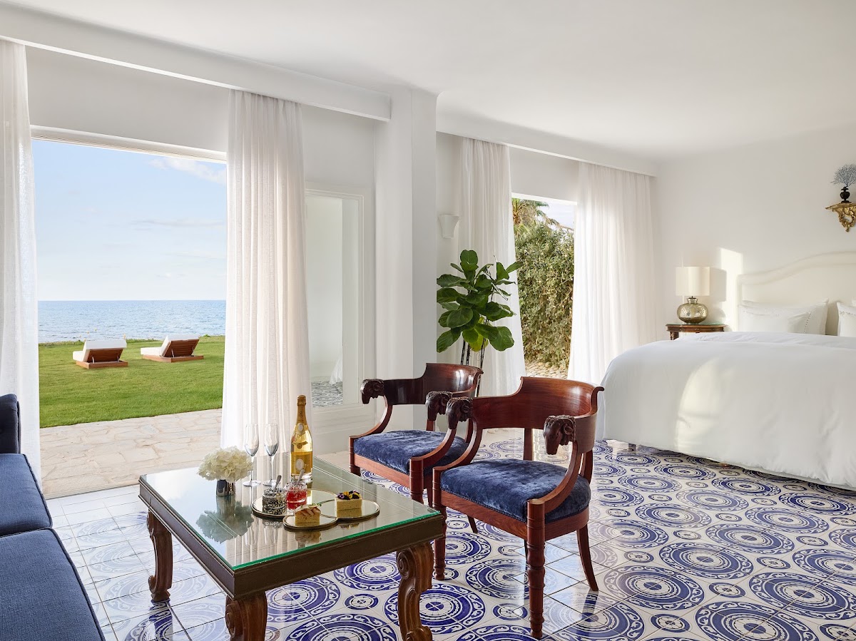 Beachfront resort suite with ocean views through open glass doors, mahogany furniture, and patterned tile flooring overlooking manicured lawn and sea.