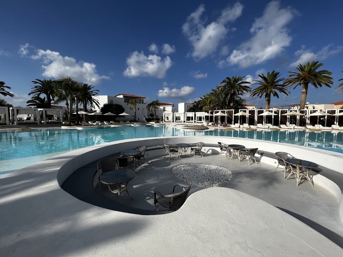 Sunken lounge area with seating and fire pit overlooking a large pool, white cabanas, and palm trees under blue sky at Grecotel Caramel Boutique Resort.