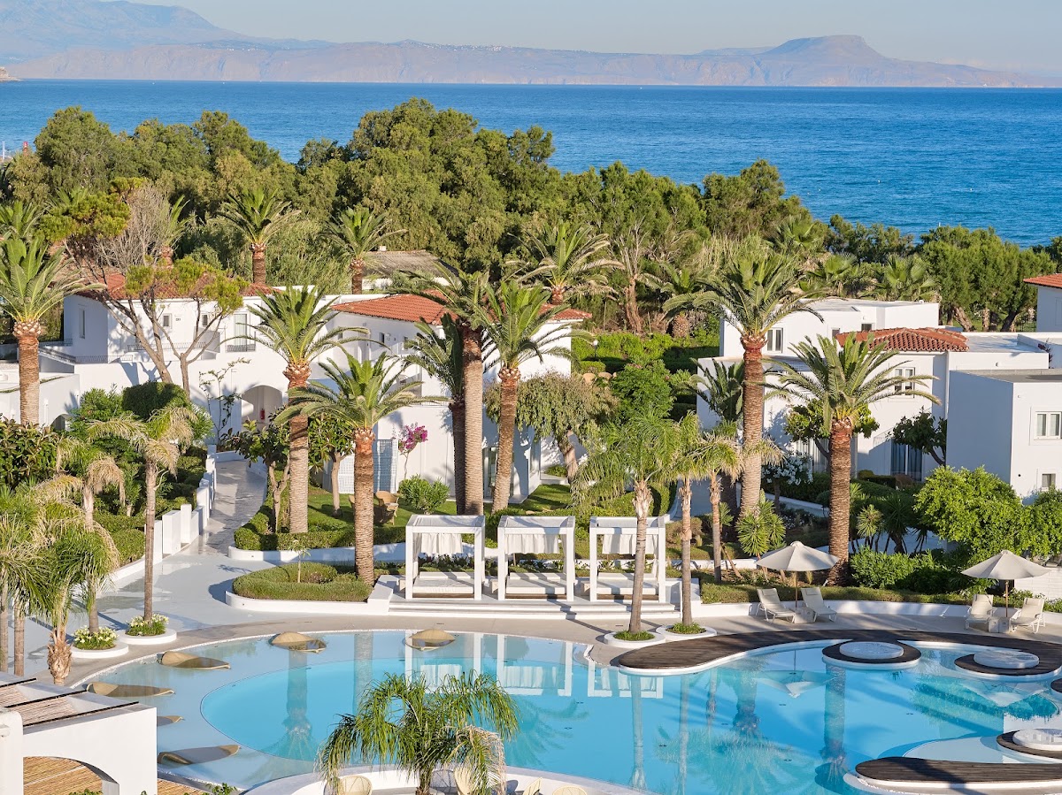 Grecotel Caramel Boutique Resort pool area with white buildings, palm trees, and mountains across the blue sea.