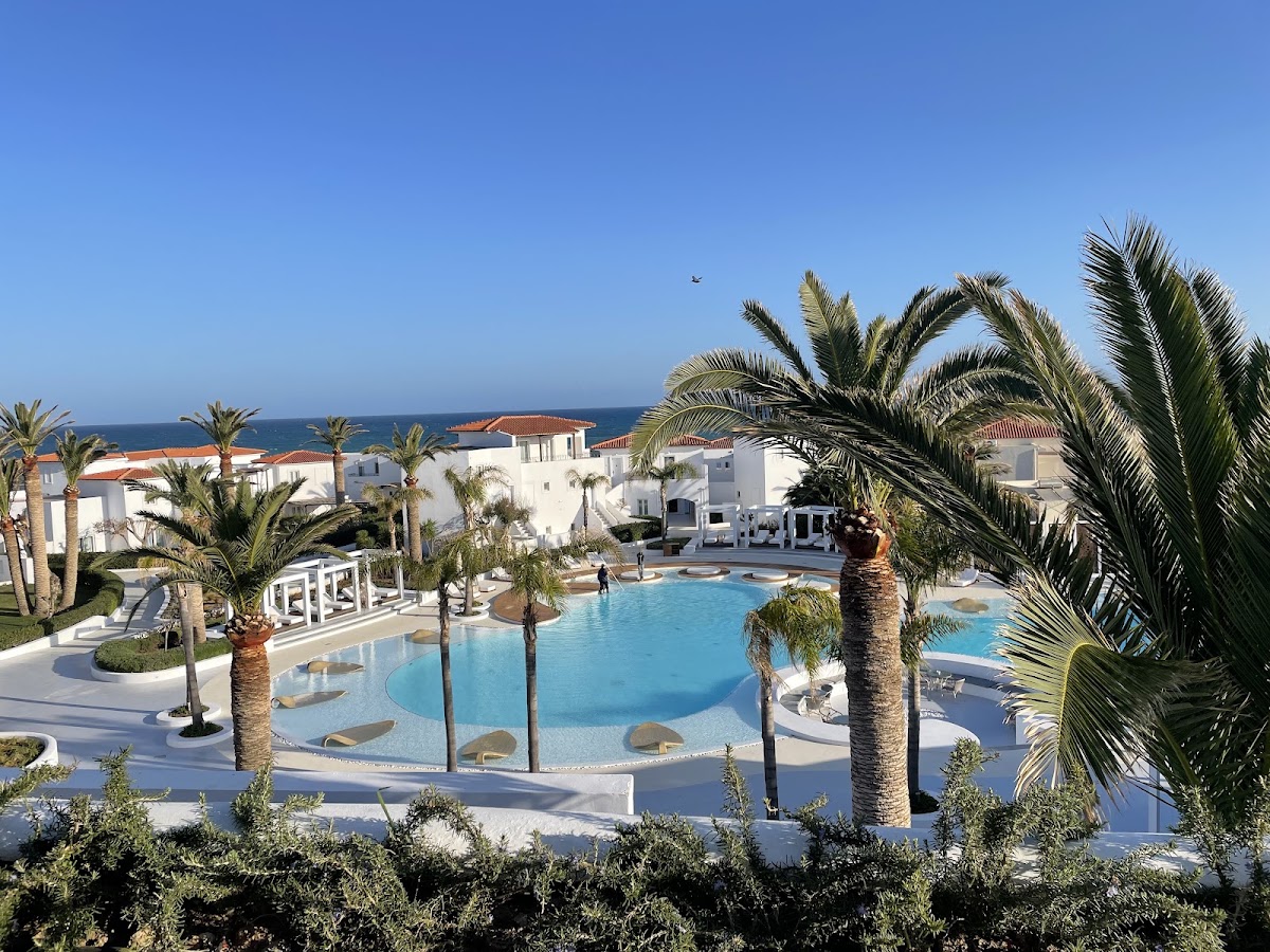 Curved turquoise pool with sunken seating surrounded by palm trees, white resort buildings, and Mediterranean Sea beyond.