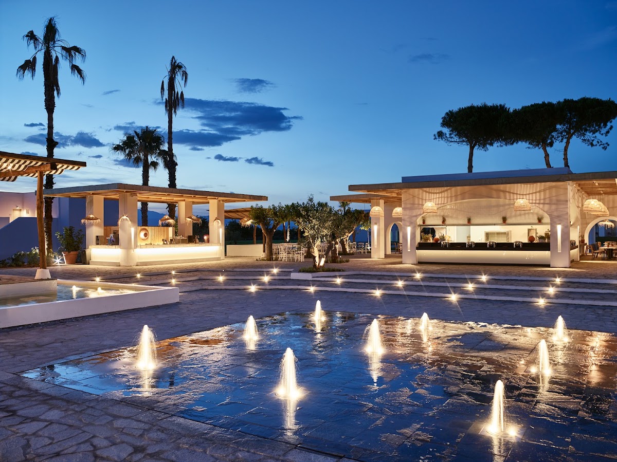 Illuminated fountains in a resort courtyard at dusk, with white pavilions, palm trees, and the sea visible in the background.