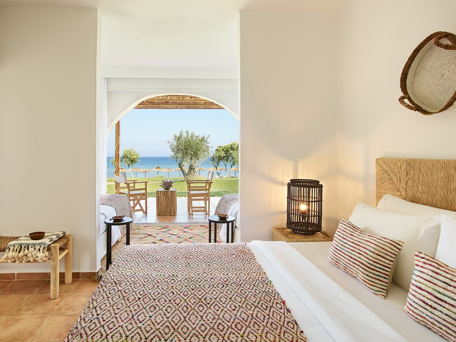 Bright bedroom with woven headboard and striped pillows opens to a terrace overlooking the beach and blue sea at Grecotel Casa Marron.