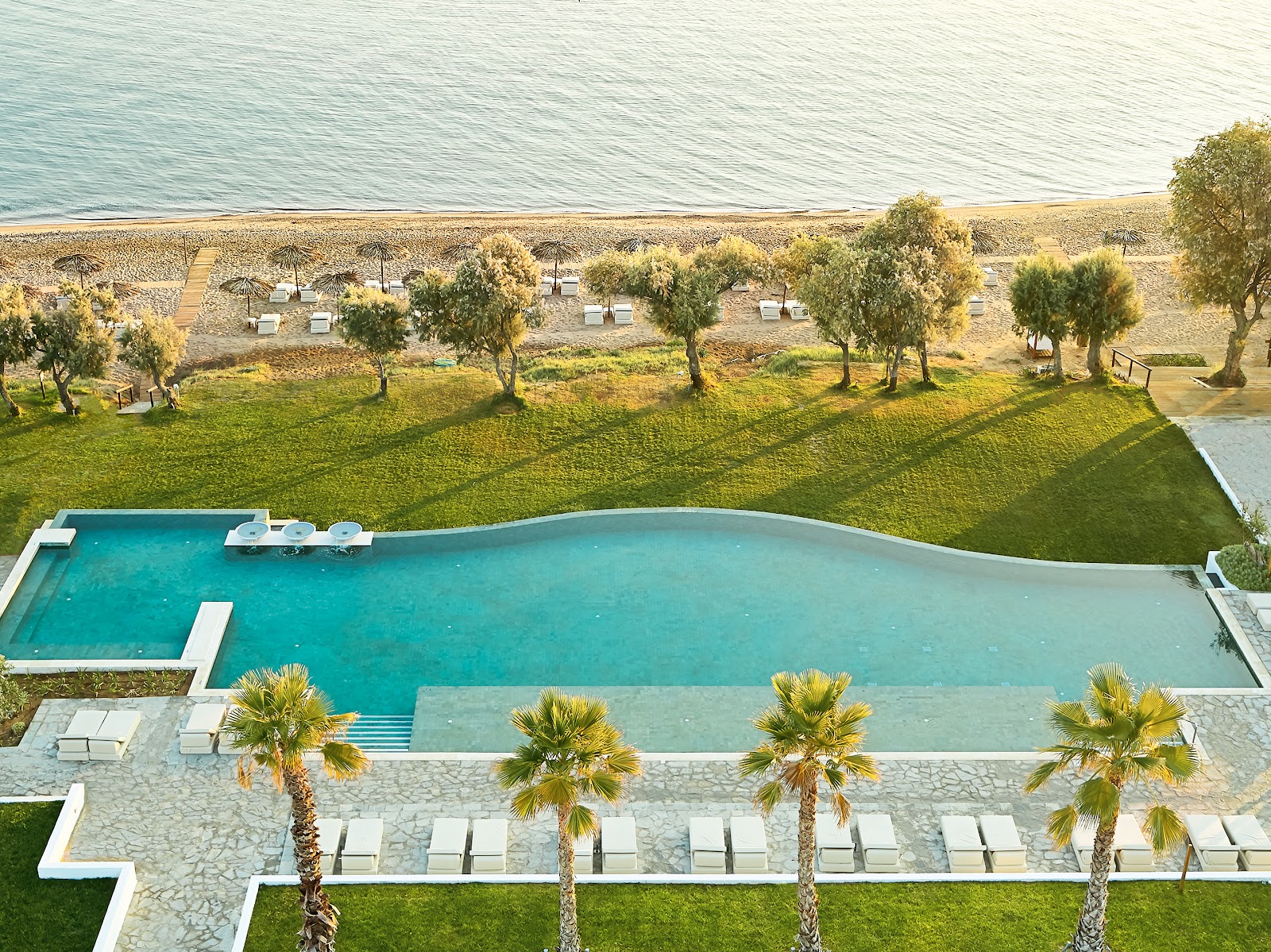 Turquoise pool with shallow end overlooking a pebble beach and calm sea, surrounded by palm trees and lounge chairs.