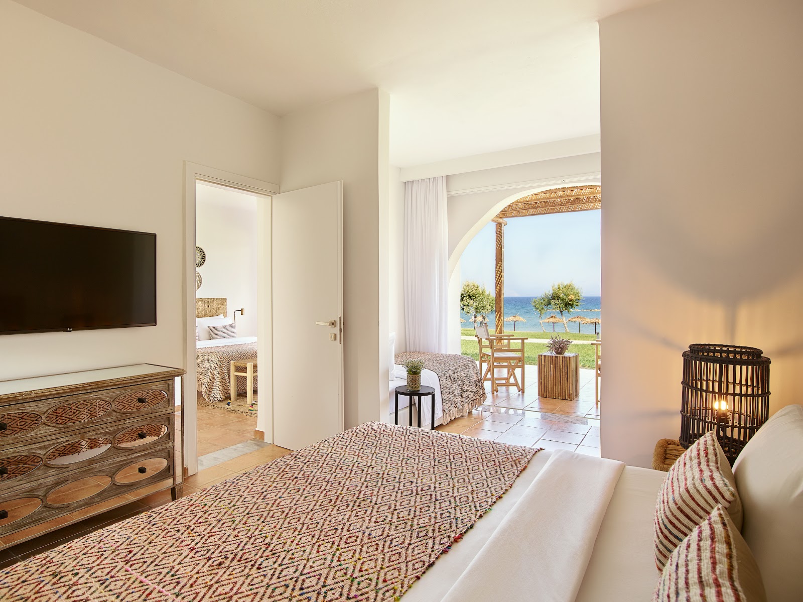Resort room with bed, TV, and open doors revealing a terrace overlooking the beach and sea at Grecotel Casa Marron.