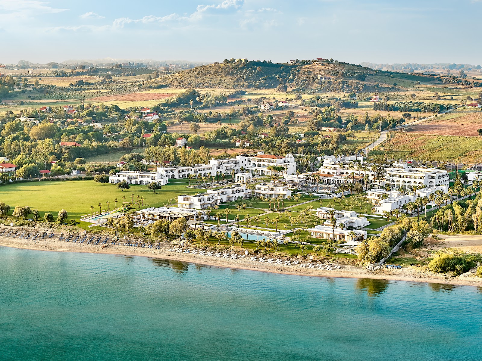 White resort buildings with pools and manicured lawns sit on a sandy beach along turquoise water, with rolling green farmland and hills rising behind.