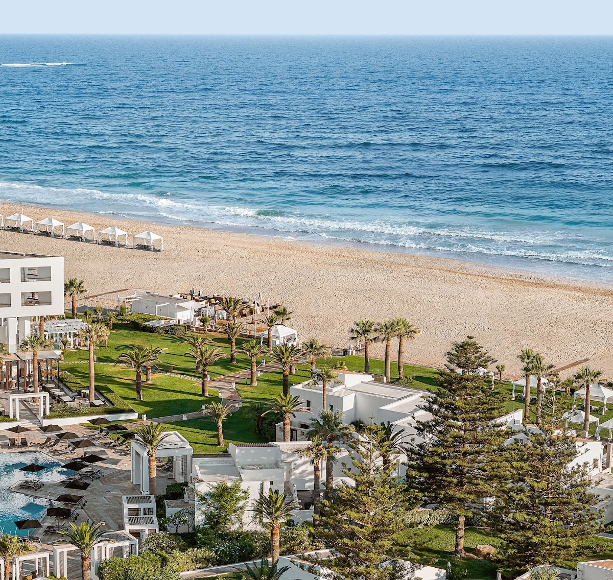 Beachfront resort grounds with white buildings, palm trees, manicured lawn, and beach cabanas overlooking the sandy beach and Mediterranean Sea.